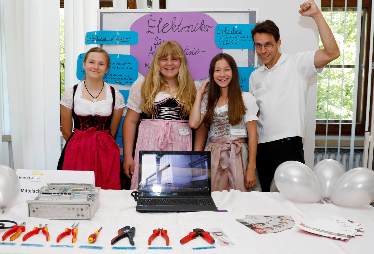 Drei Schülerinnen im Dirndl und ein Schüler stehen vor einem Stand für Elektroniker.